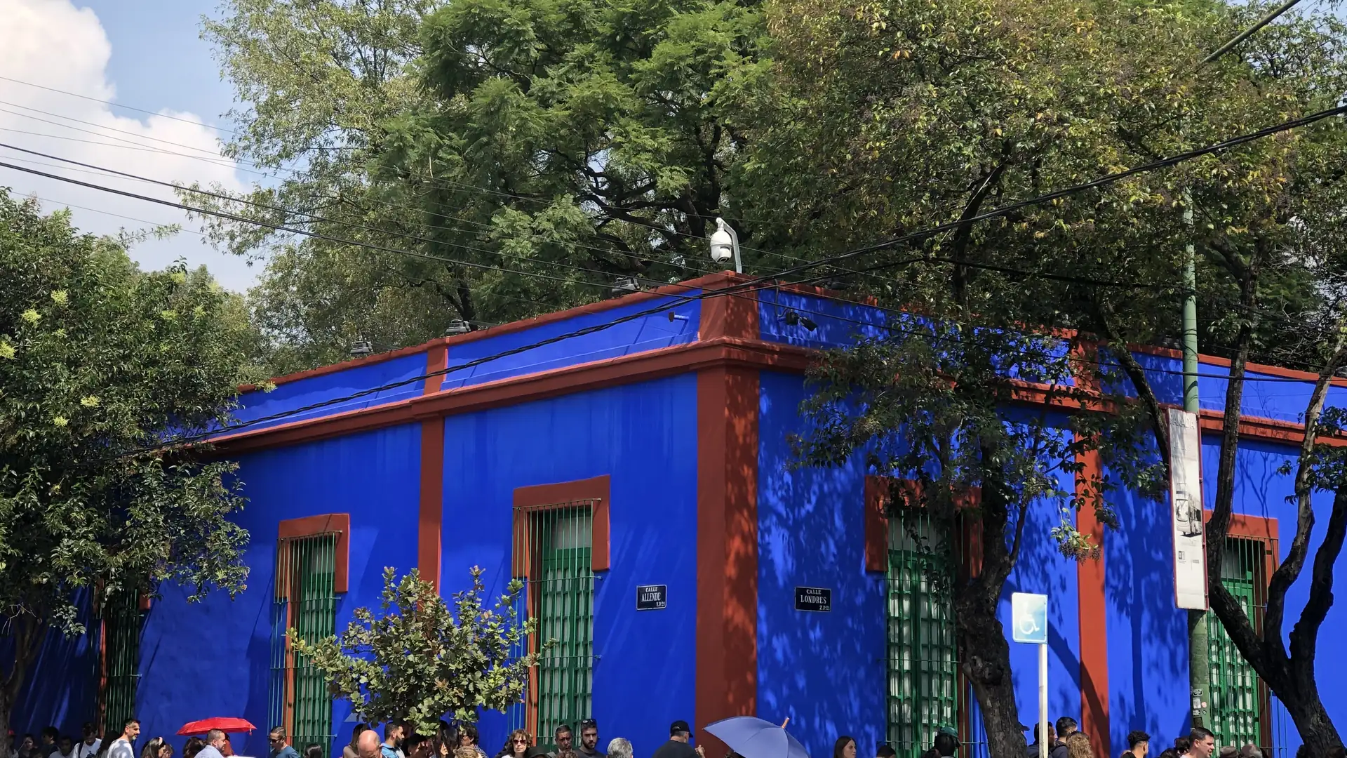 The corner of the Casa Azul in Coyoacán: deep blue walls with red-oxide trim and tall green-barred windows, under large trees and a blue sky, with visitors queuing along the pavement.