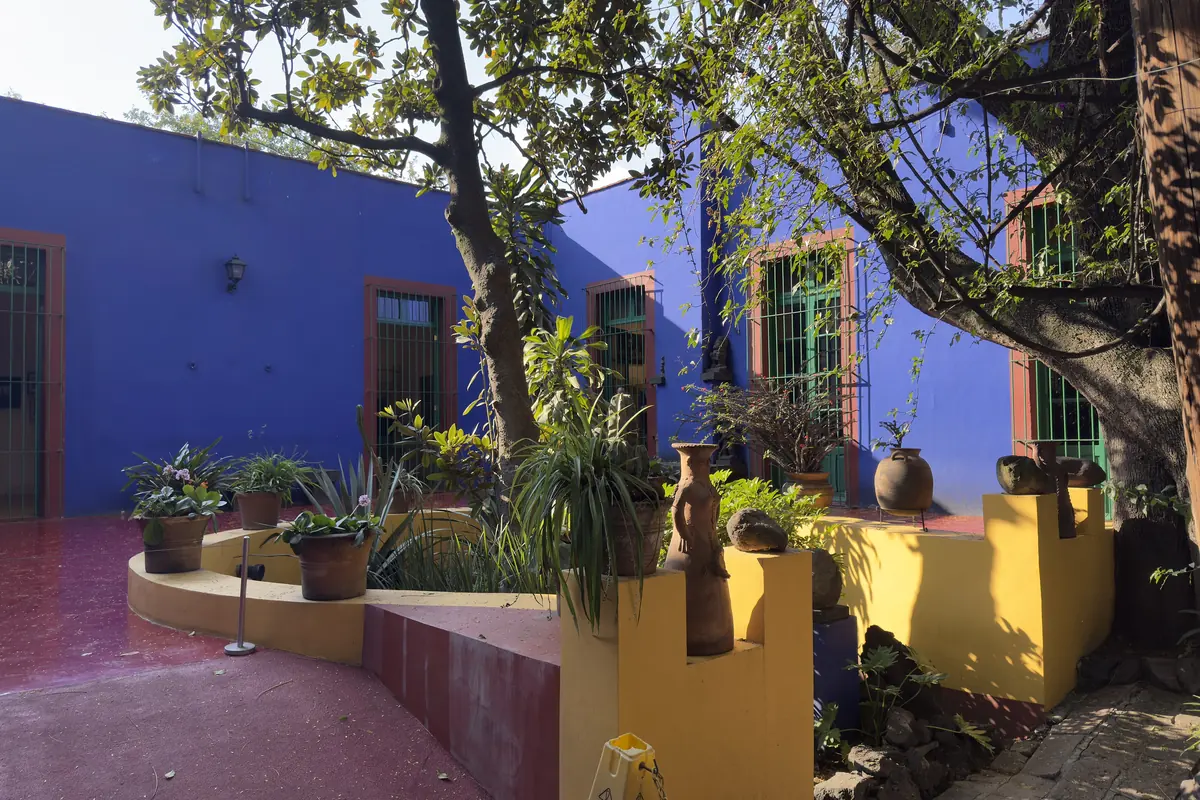 The courtyard of the Casa Azul, with deep blue walls, green window frames, potted plants on a yellow raised bed and a red-painted floor under a large tree.