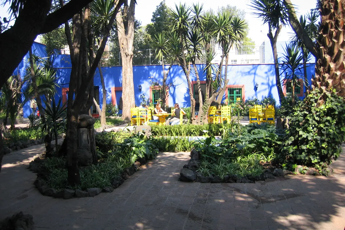 The garden of the Casa Azul: shaded stone paths between palms and trees, with yellow chairs and blue walls in the distance.