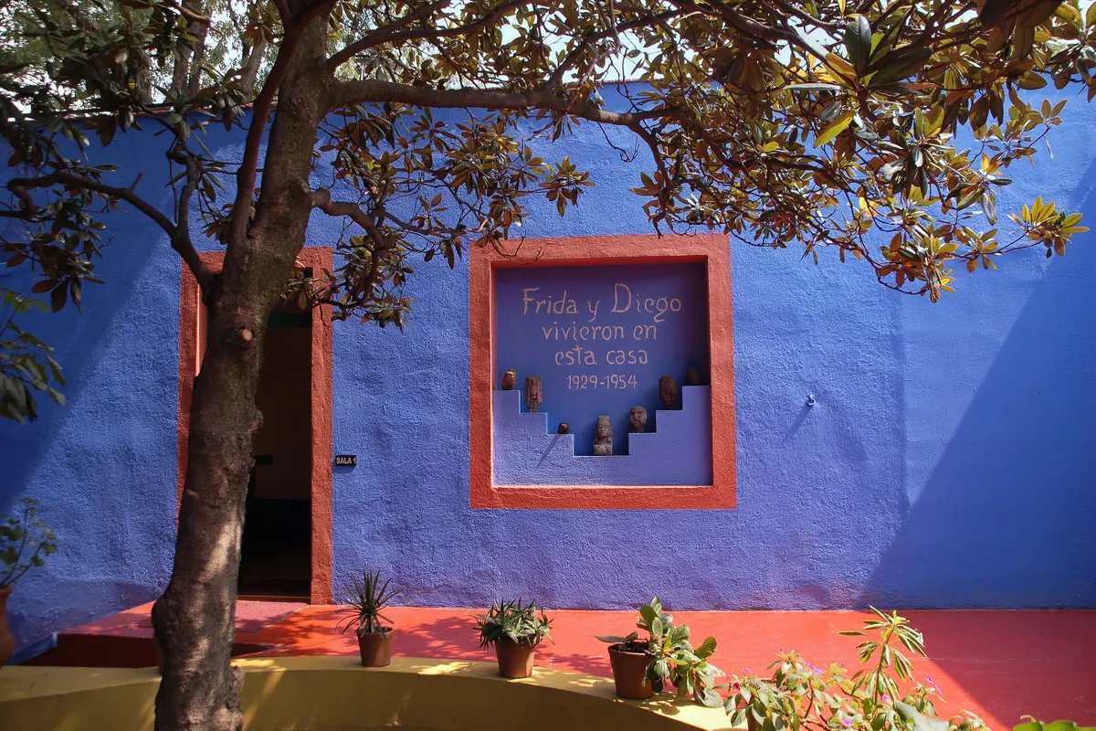 A blue courtyard wall with a red frame holding the painted words Frida y Diego vivieron en esta casa 1929–1954, above small stone figures set in stepped niches.