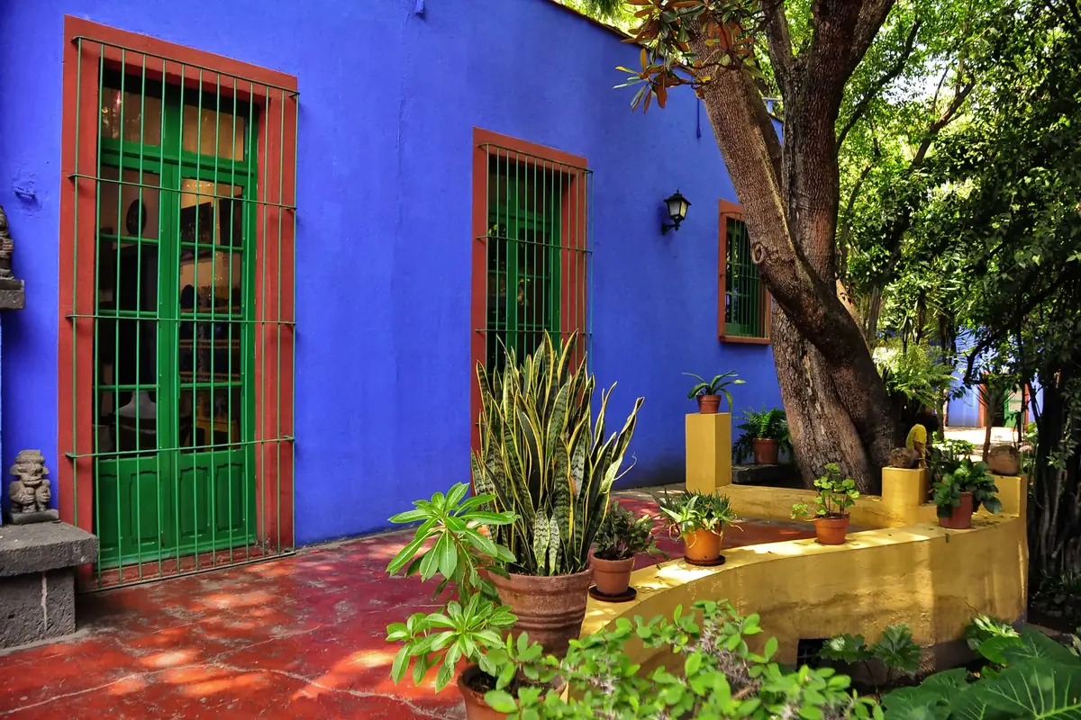 Tall green doors and windows in a blue wall with red trim, behind potted agaves and a yellow ledge in the courtyard.