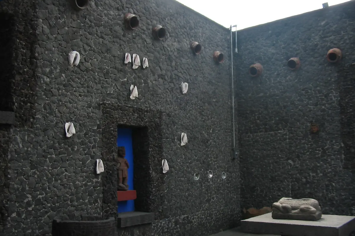 A courtyard wall of dark volcanic stone set with shells and clay pots, with a small blue niche holding a pre-Hispanic figure.